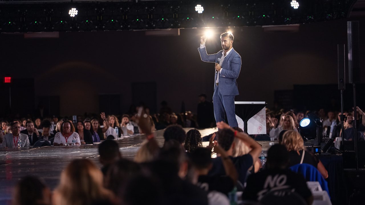 Marczell Klein under a spotlight on a wide stage, facing a packed hall.