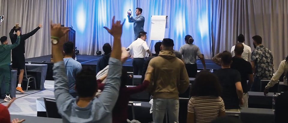 A seminar crowd with hands raised under blue stage light.