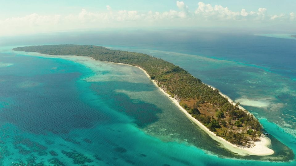 An aerial view of a long, wooded island ringed by white sand and turquoise shallows.