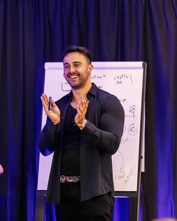 Marczell Klein on stage, smiling and applauding beside a flipchart.
