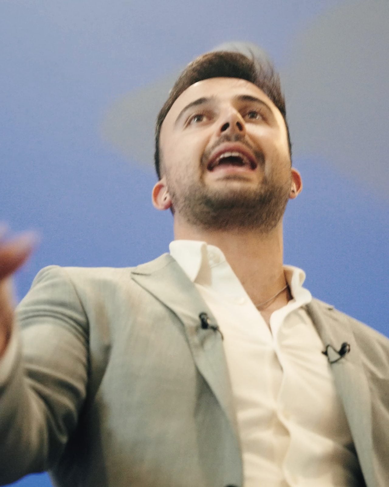 Marczell Klein on stage in a grey suit, mid-sentence, one hand reaching toward the audience.