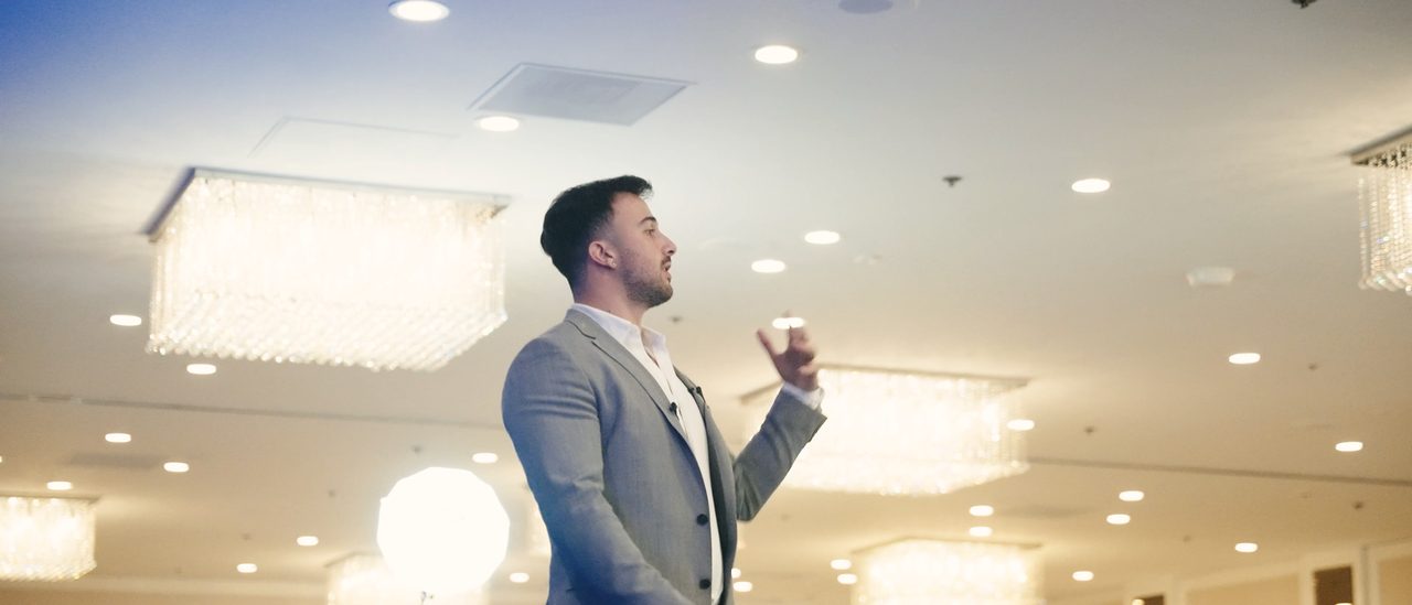 Marczell Klein on stage in a grey suit beneath the chandeliers of a hotel ballroom.