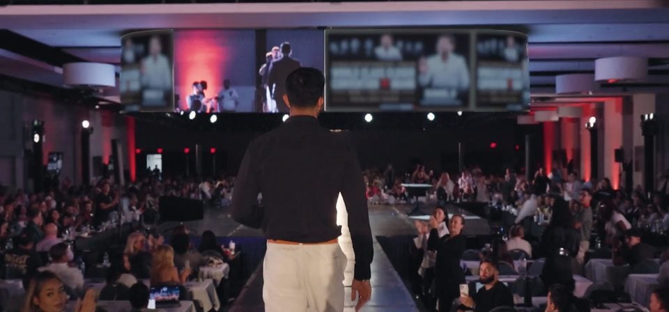 Marczell Klein walking down the runway to the stage at a live event, a packed ballroom on both sides.